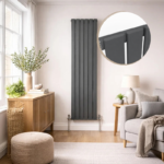 A tall, slim Luna single flat vertical designer radiator in matte anthracite grey, mounted on a light neutral wall in a modern living room next to a grey armchair and wooden sideboard.